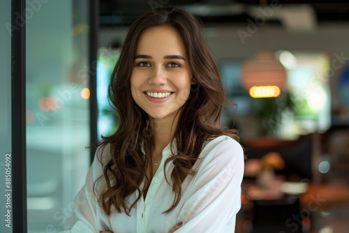 A young, beautiful brunette woman stands in a modern office, smiling happily as she looks directly at the camera, exuding confidence and professionalism.