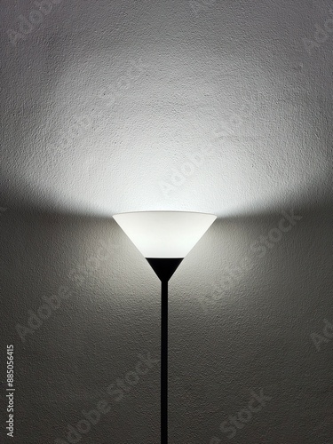 A sleek, black floor lamp with a triangular white shade is turned on, casting a soft, even light on a textured gray wall. The upward light creates a gradient effect, illuminating the area around.