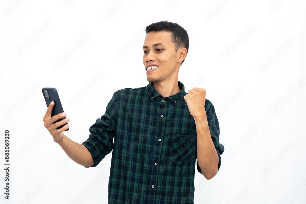Asian man wearing a green checkered casual shirt, expressing happiness and surprise while looking at a smartphone. Isolated white background. Asian man wearing a green checkered casual shirt, expressing happiness and surprise while looking at a smartphone. Isolated white background.