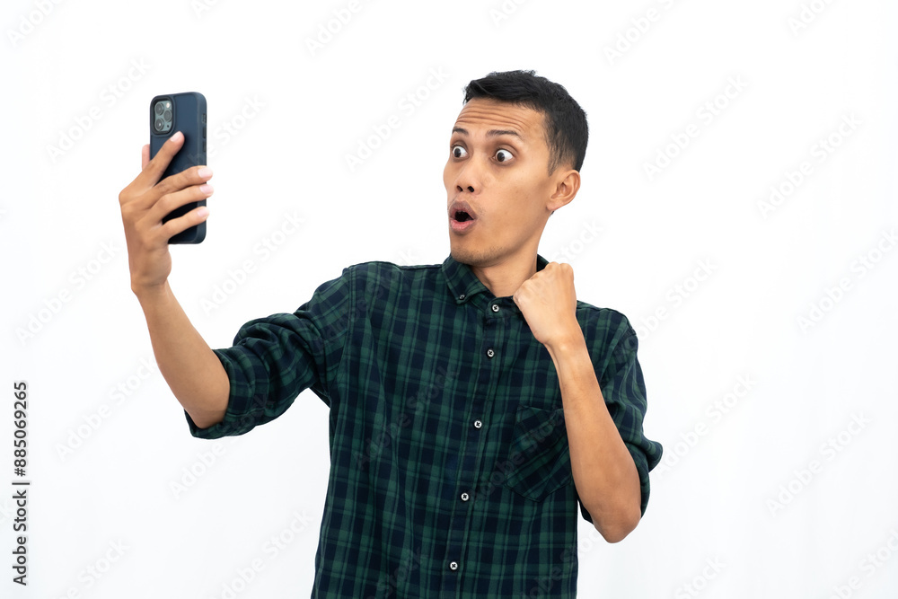 Asian man wearing a green checkered casual shirt, expressing happiness and surprise while looking at a smartphone. Isolated white background. Asian man wearing a green checkered casual shirt, expressing happiness and surprise while looking at a smartphone. Isolated white background.