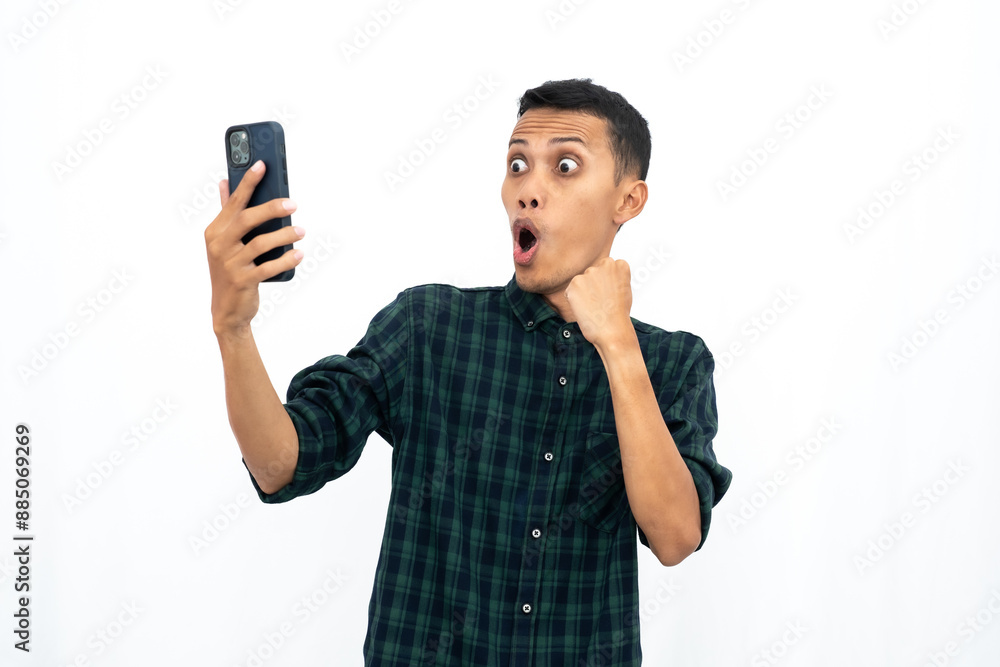 Asian man wearing a green checkered casual shirt, expressing happiness and surprise while looking at a smartphone. Isolated white background. Asian man wearing a green checkered casual shirt, expressing happiness and surprise while looking at a smartphone. Isolated white background.