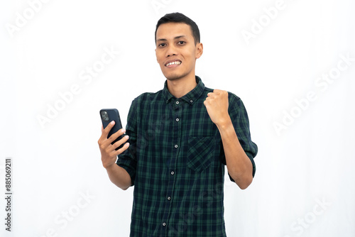 Asian man wearing a green checkered casual shirt, expressing happiness and surprise while looking at a smartphone. Isolated white background.