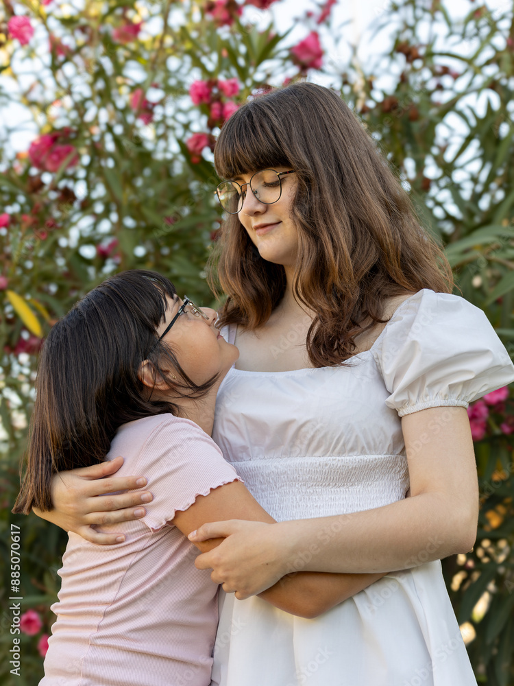 Fototapeta premium Teenage girl hugging younger sister in a garden, looking at each other with admiration