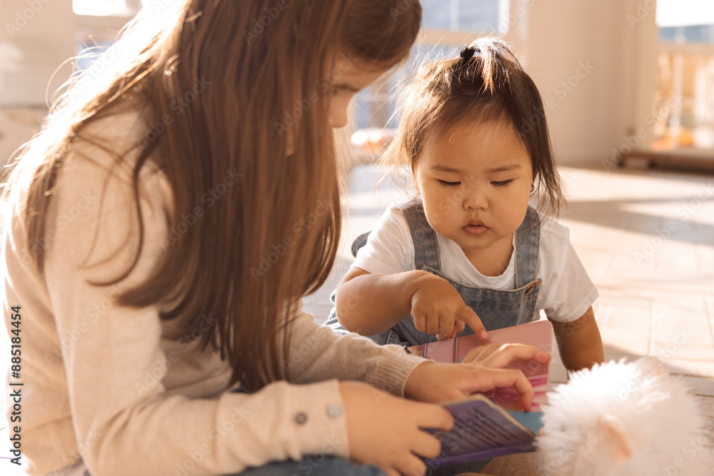 Cute children of different races play together. A Caucasian girl is reading a book to a sweet Korean baby.