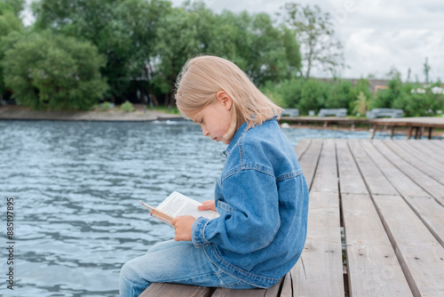 Beautiful blonde girl reads a book while sitting on the pier. Education and learning concept.