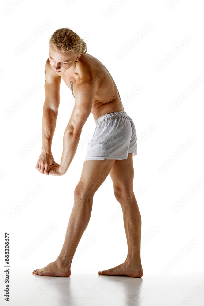 Naklejka premium Shirtless man with long blonde hair, wearing white boxer panties, clasping his hands together, showcasing his muscular arms and torso in stretching pose. Concept of natural beauty and health. Ad