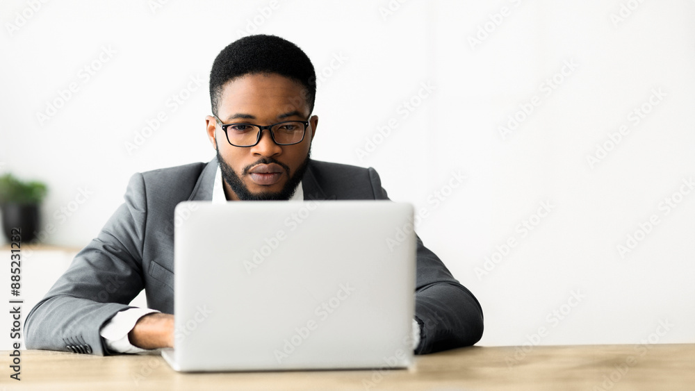 A man in a suit and glasses is working on a laptop at his desk in an office. A man in a suit and glasses is working on a laptop at his desk in an office.
