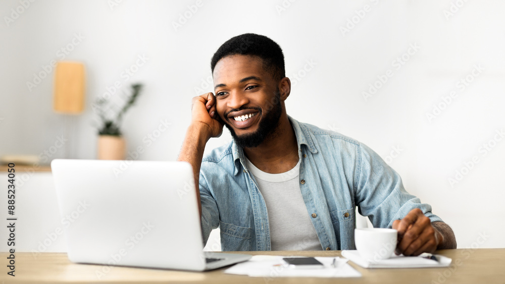 Handsome young employee talking on phone and drinking coffee while working on laptop in office, copy space Handsome young employee talking on phone and drinking coffee while working on laptop in office, copy space
