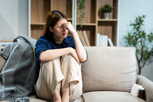 A young Asian woman sits on a sofa in her living room, feeling sick with mental health diseases. She struggles with depression, anxiety, and other conditions, seeking support and solace at home.