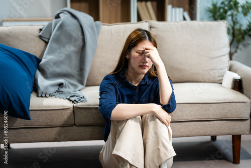 A young Asian woman sits on a sofa in her living room, feeling sick with mental health diseases. She struggles with depression, anxiety, and other conditions, seeking support and solace at home. photo