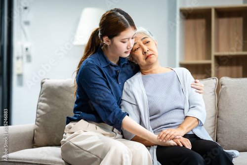 A mature mom and a young Asian woman, mother and daughter, sit together on a sofa in their living room, offering tired consolation and encouragement to each other at home.