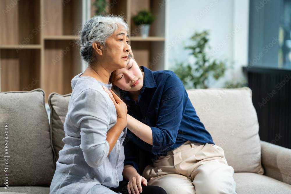 A mature mom and a young Asian woman, mother and daughter, sit together on a sofa in their living room, offering tired consolation and encouragement to each other at home. A mature mom and a young Asian woman, mother and daughter, sit together on a sofa in their living room, offering tired consolation and encouragement to each other at home.