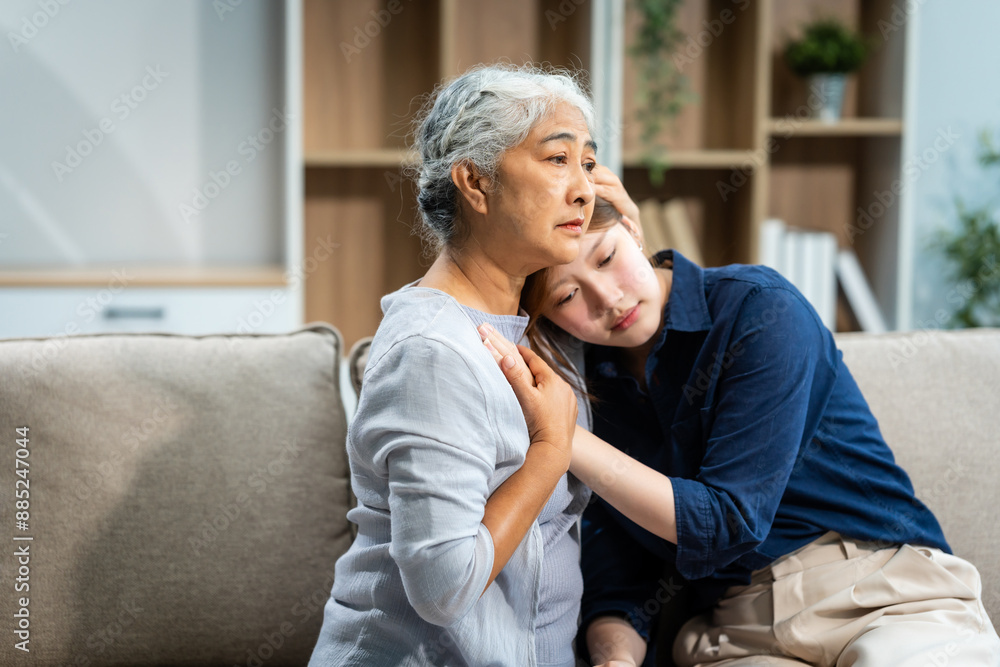 A mature mom and a young Asian woman, mother and daughter, sit together on a sofa in their living room, offering tired consolation and encouragement to each other at home. A mature mom and a young Asian woman, mother and daughter, sit together on a sofa in their living room, offering tired consolation and encouragement to each other at home.