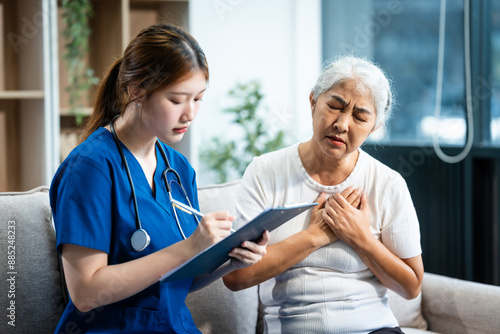 Young Asian nurse attends to older grey-haired mature woman sitting on sofa, conducting health assessment and discussing conditions osteoporosis, arthritis, Alzheimer's disease, dementia, and more.