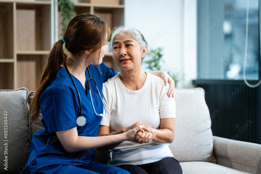 A young Asian nurse, only woman present, holds hands with an older grey-haired mature woman sitting on a sofa, offering encouragement while taking her medical history and conducting health checks. A young Asian nurse, only woman present, holds hands with an older grey-haired mature woman sitting on a sofa, offering encouragement while taking her medical history and conducting health checks.