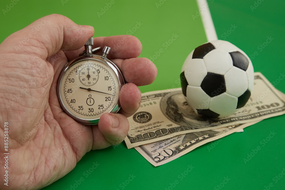 Time on stopwatch in hand and dollars with footbal ball on green background, betting time, betting acceptance, betting winnings .. Time on stopwatch in hand and dollars with footbal ball on green background, betting time, betting acceptance, betting winnings ..