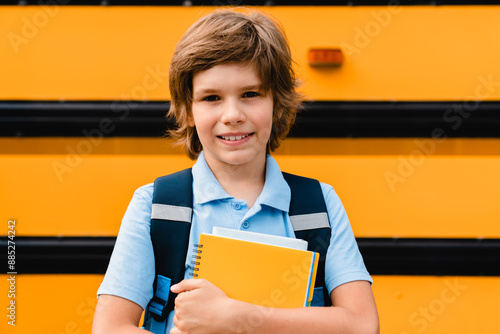 Small kid young student pupil boy coming back to school after summer holidays waiting next to the school bus holding notebooks and copybooks preparing for new semester educational year photo