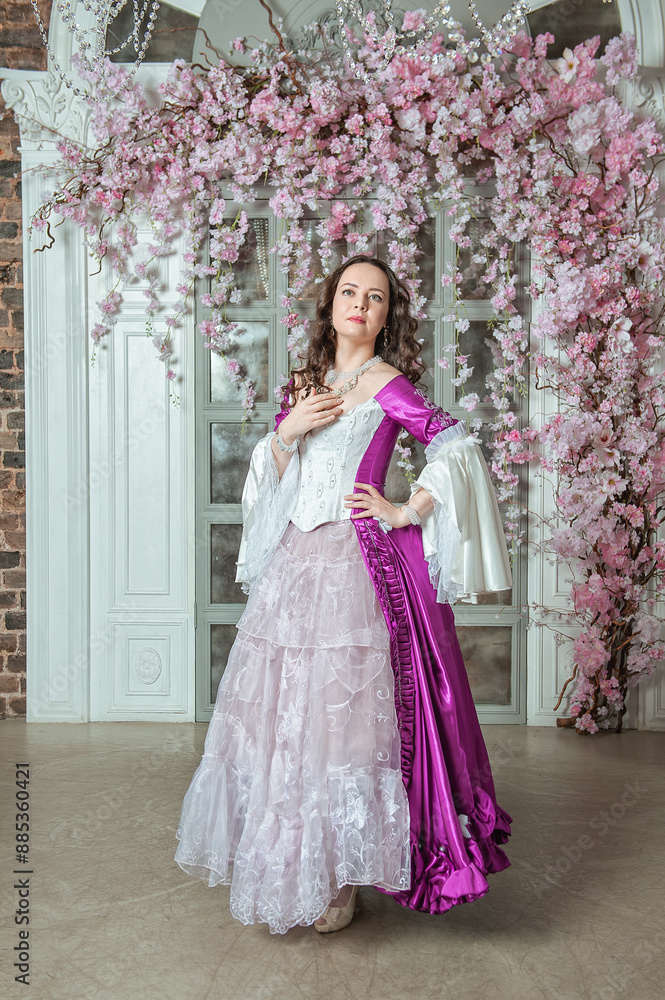 Beautiful woman in fantasy white and purple rococo style medieval dress standing near wall with pink flowers