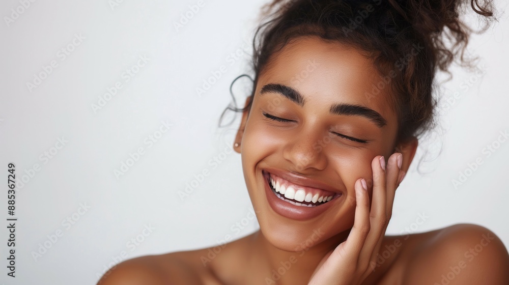 Model looking young ethnic young woman playful laugh with glowing skin and perfect teeth open shoulders and eyes closed on white background with hand on her cheek and hair bun Model looking young ethnic young woman playful laugh with glowing skin and perfect teeth open shoulders and eyes closed on white background with hand on her cheek and hair bun