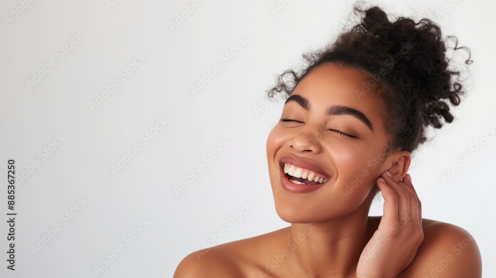Model looking young ethnic young woman playful laugh with glowing skin and perfect teeth open shoulders and eyes closed on white background with hand on her cheek and hair bun Model looking young ethnic young woman playful laugh with glowing skin and perfect teeth open shoulders and eyes closed on white background with hand on her cheek and hair bun