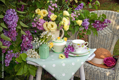 Still life of lilac and Eustoma in a jug on a table with teacup saucer and plate on a wooden table with wicker chair in a garden with lilac tree. 