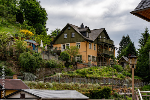 Wallpaper Mural A house with a green roof sits on a hillside. The house is surrounded by a lush green garden and has a large patio area. The sky is cloudy, giving the scene a somewhat moody atmosphere Torontodigital.ca