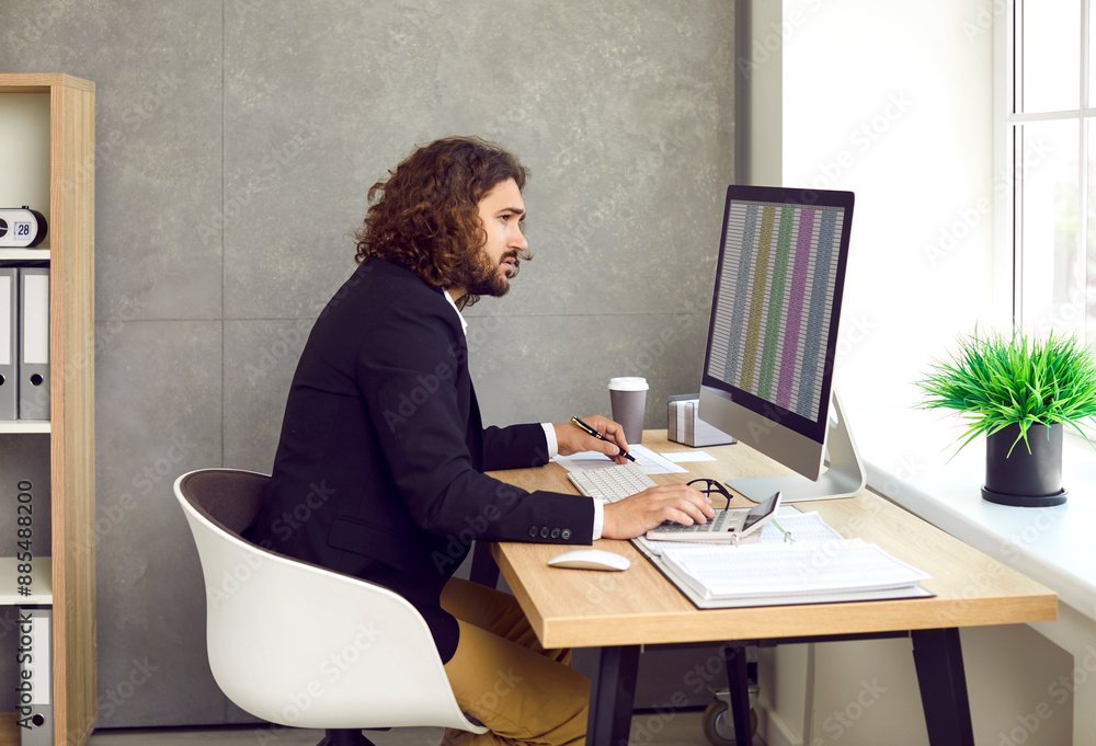 Side view of a young stylish bearded business man working on a laptop with tables and charts at the office or at home and making calculations. Business accounting, data analysis and finance concept. Side view of a young stylish bearded business man working on a laptop with tables and charts at the office or at home and making calculations. Business accounting, data analysis and finance concept.