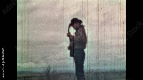 Man playing guitar, singing on cloudy sky background in wild field nature. Male cowboy in hat enjoy playing musical instrument in summer outdoors. Vintage color film. Travel archive. Retro 1960s