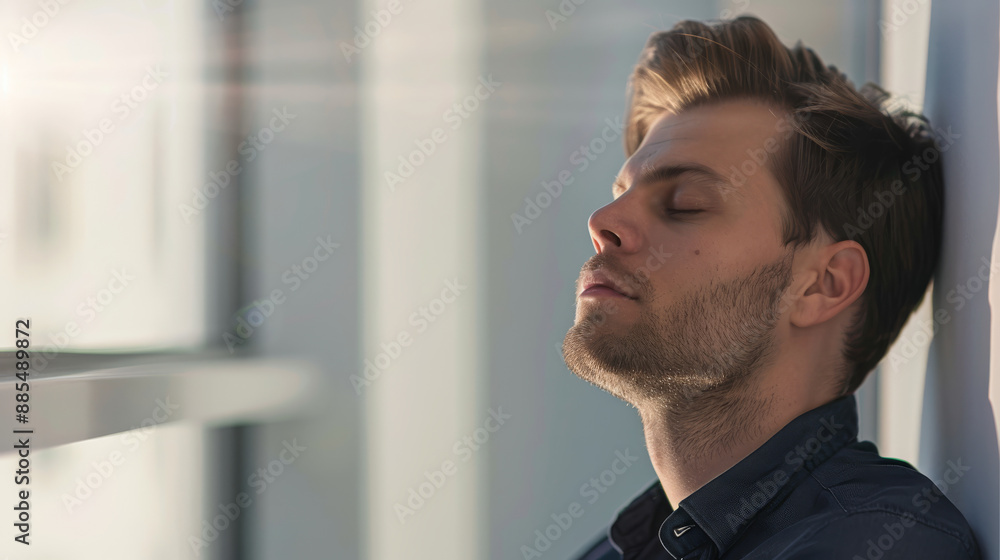 Naklejka premium A contemplative man with closed eyes leans against a window, basking in soft afternoon light, exuding a calm, reflective mood.