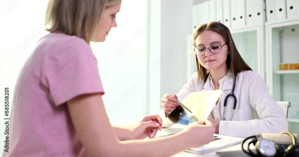 Serious physician shows female patient x-ray image of damaged wrist sitting at table in hospital. Attentive woman nods looking at radiograph picture slow motion Serious physician shows female patient x-ray image of damaged wrist sitting at table in hospital. Attentive woman nods looking at radiograph picture slow motion