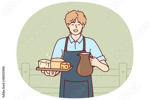 Man holds jug of milk and tray of cheese from cow farm selling own organic food. Guy wearing apron working on cow farm offers to buy organic dairy products with useful nutritional vitamins.