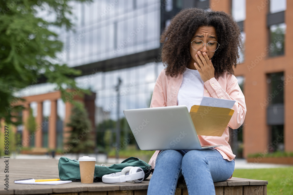 Woman student looking worried while reading letter on laptop outdoors at university campus. Concept of education, stress, university life, bad news, financial issues. Woman student looking worried while reading letter on laptop outdoors at university campus. Concept of education, stress, university life, bad news, financial issues.