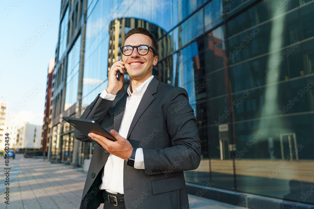 A smiling businessman, holding a tablet and talking on the phone, standing in a modern urban area. A smiling businessman, holding a tablet and talking on the phone, standing in a modern urban area.