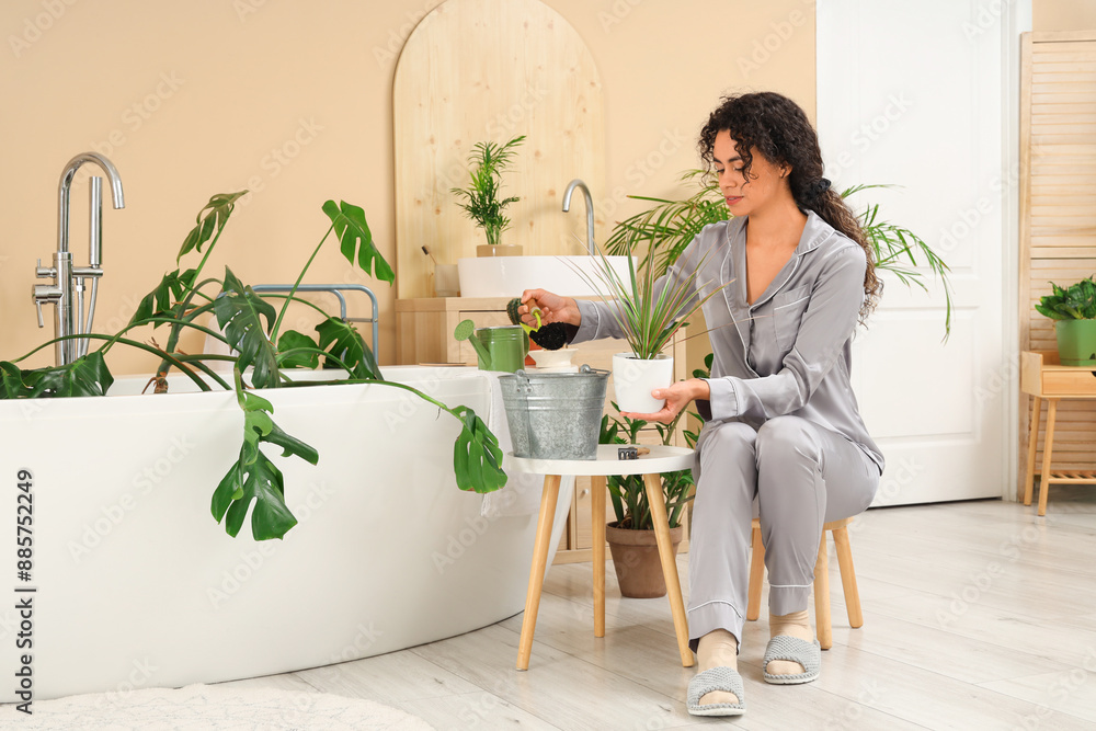 Young African-American woman putting soil into pot with plant in bathroom Young African-American woman putting soil into pot with plant in bathroom
