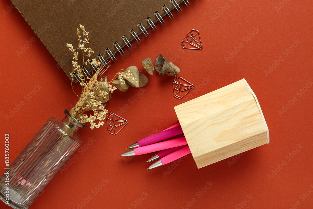 Vase of dried flowers, wooden holder with pens and notebook on red background Vase of dried flowers, wooden holder with pens and notebook on red background