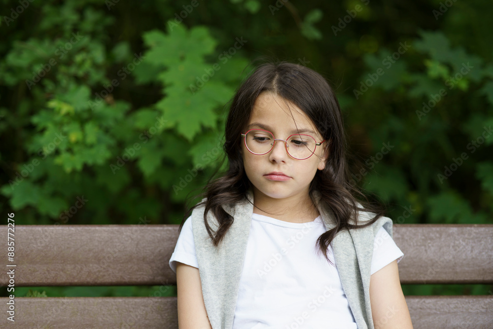 A little sad schoolgirl girl with glasses sits on a bench. Difficult childhood concept A little sad schoolgirl girl with glasses sits on a bench. Difficult childhood concept