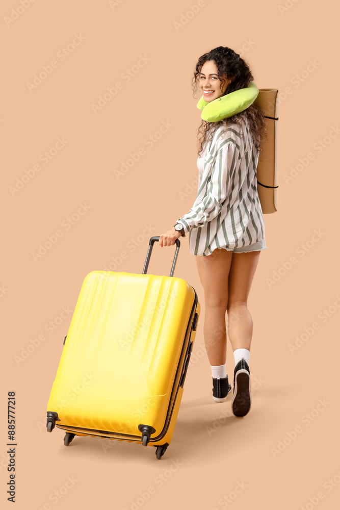 Young African-American woman with neck pillow and baggage going somewhere on beige background Young African-American woman with neck pillow and baggage going somewhere on beige background