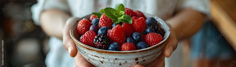 Hand holding a bowl filled with fresh berries, including strawberries, blueberries, and raspberries, garnished with mint leaves. Hand holding a bowl filled with fresh berries, including strawberries, blueberries, and raspberries, garnished with mint leaves.