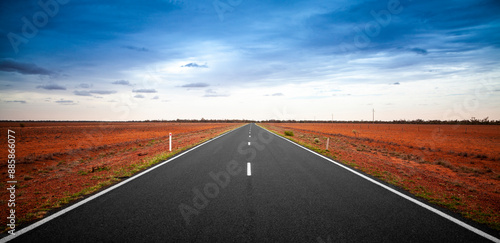 Outback Queensland road in Australia with a stormy sky. An empty open road stretching to the horizon. Single-lane sealed highway with deep orange-red soil on both sides. Travel adventure.