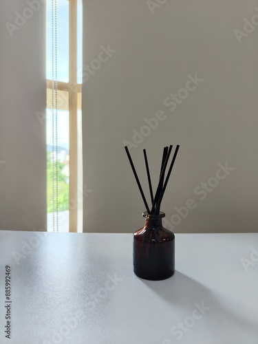A minimalist setup with a small, dark glass perfume bottle holding several black reed sticks sits on a light-colored surface. In the background, soft light filters through partially covered windows. 
