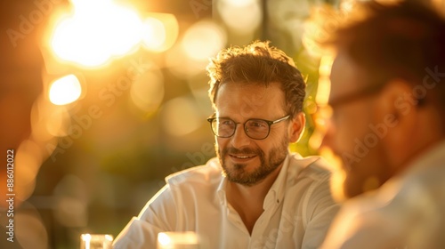 Smiling man with glasses enjoying an outdoor gathering with friends during sunset, creating a warm and cheerful atmosphere.