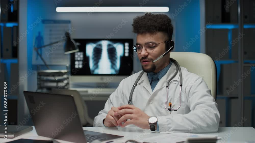 Medium shot of bearded doctor in glasses, lab coat, with stethoscope and call center set speaking with patient on laptop, having online appointment in modern clinic Medium shot of bearded doctor in glasses, lab coat, with stethoscope and call center set speaking with patient on laptop, having online appointment in modern clinic