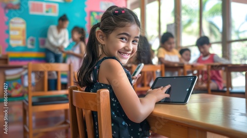 Girl at a desk with an iPad, smiling, in a colorful classroom with other students and a teacher.