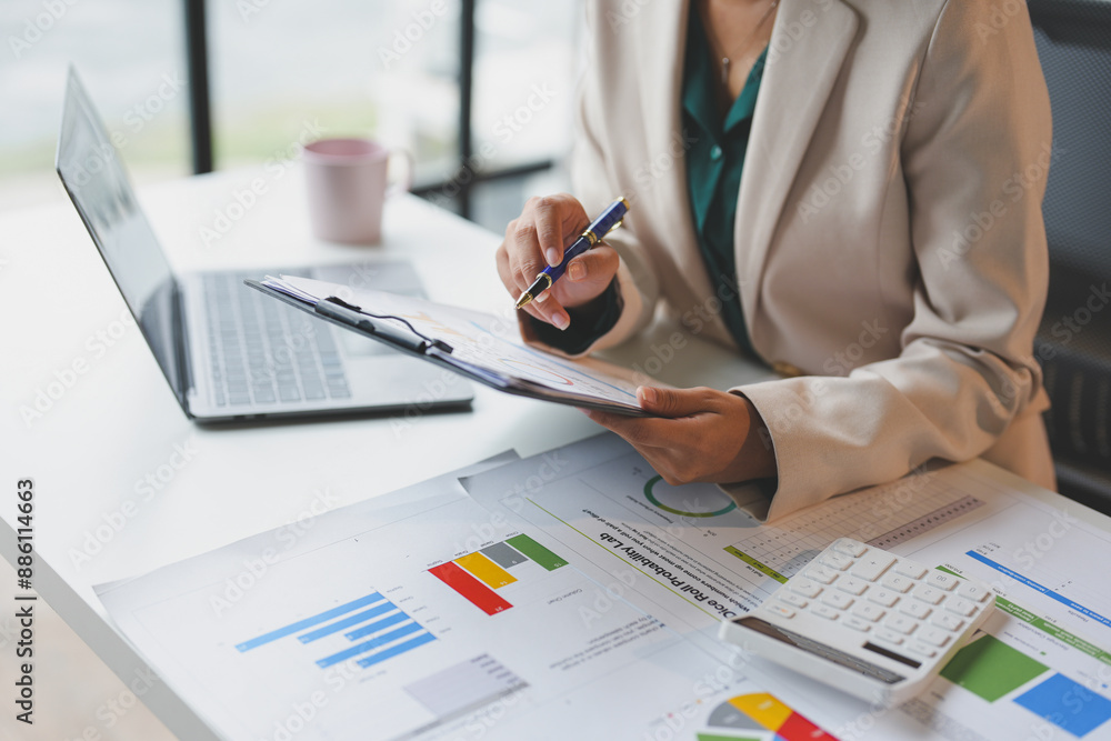 Naklejka premium Happy Asian businesswoman in office working on financial documents, holding pen pointing at laptop. Results, income, online marketing. Company taxes Online business work.