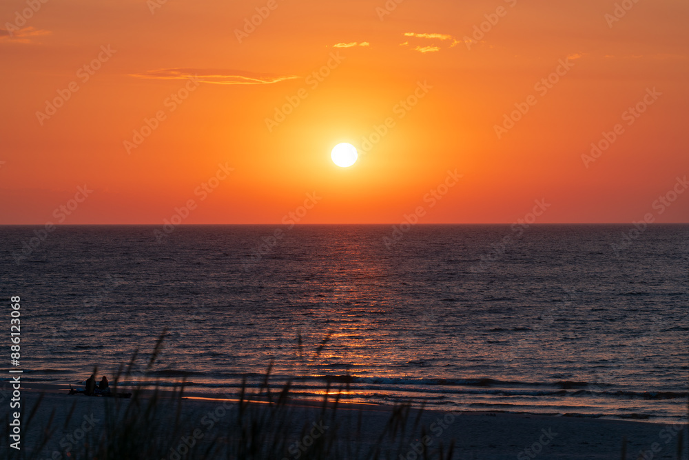 Beautiful sunset over the Baltic Sea. Blurry blades of grass in the foreground. Silhouettes of two people sitting on a beautiful wide beach and watching the sunset. Warm orange sky. Slajszewo, Poland