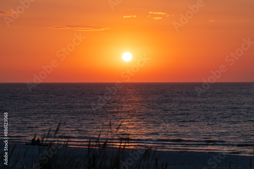 Obraz na płótnie Beautiful sunset over the Baltic Sea. Blurry blades of grass in the foreground. Silhouettes of two people sitting on a beautiful wide beach and watching the sunset. Warm orange sky. Slajszewo, Poland w salonie