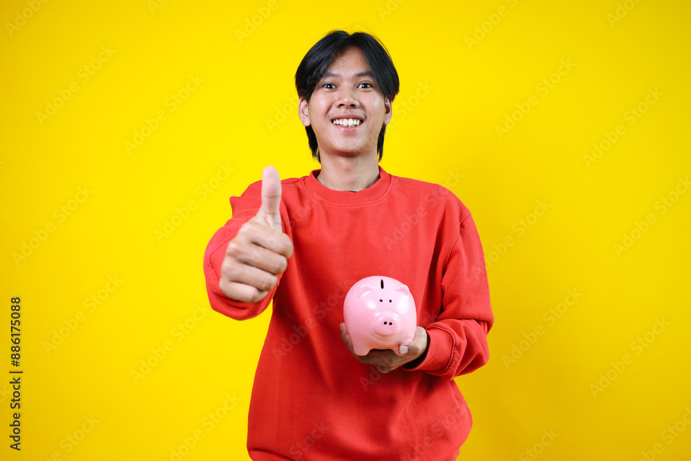 Young Asian man holding piggy bank for savings while showing thumb up isolated over yellow background Young Asian man holding piggy bank for savings while showing thumb up isolated over yellow background