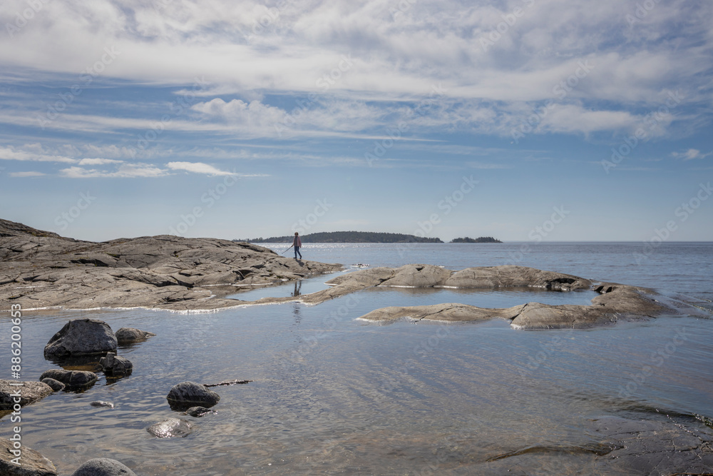 Fototapeta premium A person strolls on rocks by a serene lake. Clear blue water mirrors the sky above, with far-off shoreline and islands barely seen through haze.