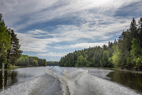 A tranquil view of a Finnish lake on a summer day. The water is calm and clear, reflecting the blue sky and white clouds above. The shores are lined with lush green trees.