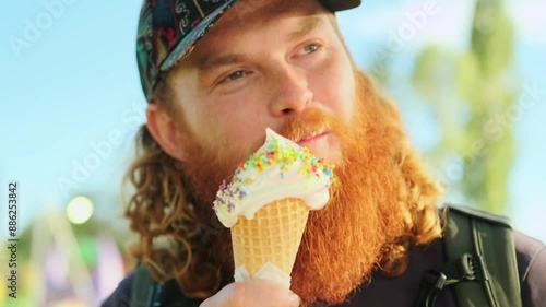 A red-haired curly guy in love eats ice cream with multi-colored sprinkles on a waffle cone, dirtying his beard and smiling in summer weather, close up.